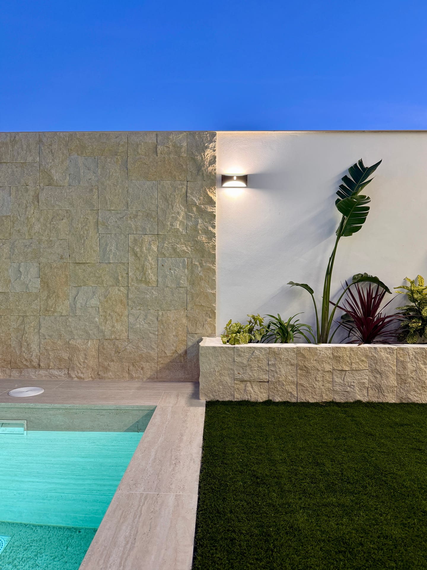 Piscina, césped y jardinera con plantas tropicales junto al muro de piedra